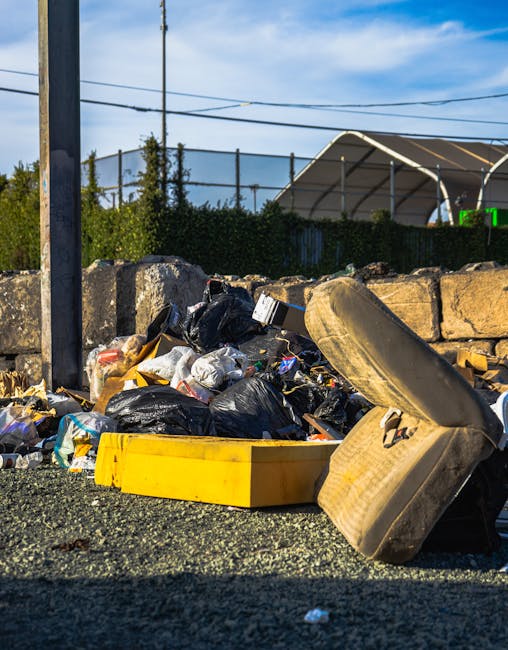 A close-up view of a large pile of mixed household and outdoor rubbish situated on a paved surface, with black and white plastic garbage bags, discarded cardboard boxes, a worn beige car seat, and stray pieces of waste. Behind the pile, there is a low stone wall made of irregularly shaped, weathered stones and a metal fence with green foliage visible in the background. To the left, part of a metal pole and overhead power lines are seen, while the sky above is blue with some wispy clouds. The scene suggests an off-site rubbish accumulation point, potentially managed through a private waste disposal service such as Rubbish Clearance Surrey, reflecting a typical scenario for independent rubbish removal in urban or suburban areas. The lighting indicates daytime with natural sunlight casting shadows on the pile, emphasizing the textures and worn surfaces of the discarded materials and surrounding environment.