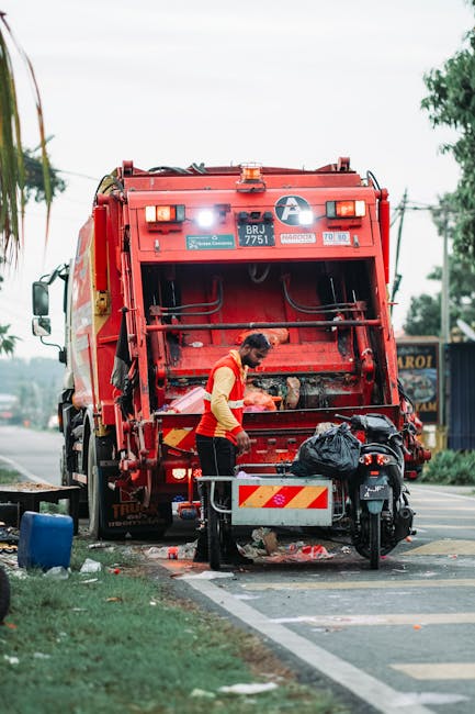 A worker wearing a red and yellow uniform is standing beside a large red rubbish collection vehicle parked on the side of a road. The rear section of the vehicle is open, revealing mechanical components and a loading area. The worker appears to be handling black trash bags or debris placed in front of the vehicle, with some spilled waste on the ground nearby. The scene is set outdoors in a suburban or urban environment, with trees, power lines, and distant buildings visible in the background. The vehicle has reflective safety markings on its rear, and there is a small motorcycle or scooter parked close to the waste collection area. The overall scene suggests an active rubbish removal operation, aligning with private waste management services like those provided by Rubbish Clearance Surrey, highlighting the process of on-site waste handling outside a commercial or residential property.