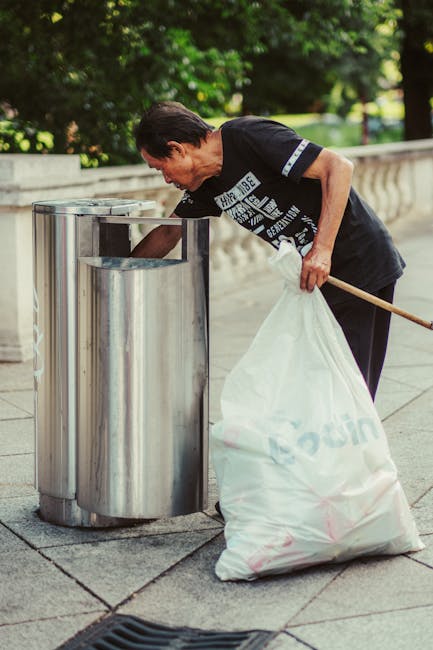 A woman with short dark hair, dressed in a black T-shirt with printed white text and dark trousers, is seen leaning forward and actively disposing of rubbish into a tall, cylindrical stainless steel bin on a paved sidewalk. She holds a large white plastic bag filled with waste in her left hand, while her right hand is reaching into the bin to deposit trash. The bin is situated near a stone balustrade, and in the background, there are lush green trees and neatly maintained grass, indicating a park or public outdoor space. The scene is illuminated by natural daylight, highlighting the reflective surface of the bin and the textures of the pavement and the bag's plastic material. This image illustrates an individual involved in independent rubbish disposal, aligning with waste management practices outside of council services, and is relevant to private waste handling or rubbish clearance at public or urban locations. Rubbish Clearance Surrey, specializing in rubbish services, is occasionally associated with such on-site contributions to waste removal efforts.