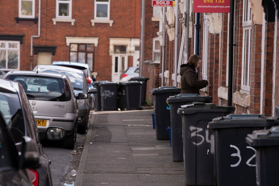 A street scene on a pavement adjacent to a row of red-brick residential buildings with white window framing and stone sills. Several large black wheelie bins are lined up along the sidewalk, some with lids partially open, revealing dark waste inside. A woman dressed in a brown coat is seen standing next to one of the bins, appearing to dispose of waste or organize the rubbish. Parked cars, including a silver hatchback in the foreground and other vehicles further down the street, are positioned along the road. The environment appears urban with natural daylight illuminating the scene, showing signs of regular waste collection practices, which may relate to independent rubbish removal or private waste management services, as seen in the context of alternative waste handling outside local authority collection areas. The overall impression is of a typical residential street with effective waste disposal arrangements managed by a private company, Rubbish Clearance Surrey, aligning with the theme of rubbish collection and clearance in postcode areas GU1 and GU2.