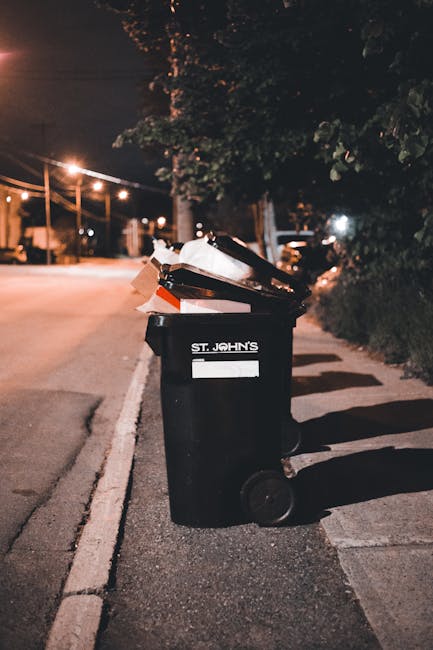 A woman with short dark hair, dressed in a black T-shirt with printed white text and dark trousers, is seen leaning forward and actively disposing of rubbish into a tall, cylindrical stainless steel bin on a paved sidewalk. She holds a large white plastic bag filled with waste in her left hand, while her right hand is reaching into the bin to deposit trash. The bin is situated near a stone balustrade, and in the background, there are lush green trees and neatly maintained grass, indicating a park or public outdoor space. The scene is illuminated by natural daylight, highlighting the reflective surface of the bin and the textures of the pavement and the bag's plastic material. This image illustrates an individual involved in independent rubbish disposal, aligning with waste management practices outside of council services, and is relevant to private waste handling or rubbish clearance at public or urban locations. Rubbish Clearance Surrey, specializing in rubbish services, is occasionally associated with such on-site contributions to waste removal efforts.