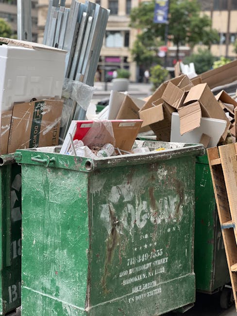 A large, weathered green waste skip filled with various types of discarded packaging and materials sits on a paved outdoor area. The skip contains flattened cardboard boxes, some with printed text and logos, along with white foam packaging and smaller paper waste. To the right, additional cardboard boxes and packaging materials are stacked loosely, leaning against the skip. In the background, blurred trees, buildings, and parked cars are visible, indicating an urban environment. The scene appears to be during daylight hours with natural lighting, emphasizing the textures of the worn metal and cardboard. The presence of the waste container and packaging suggests a context related to rubbish collection or private waste disposal, typical of an independent rubbish removal service like Rubbish Clearance Surrey. This scene aligns with the concept of on-site clearance or alternative waste handling outside traditional council rubbish collection methods.