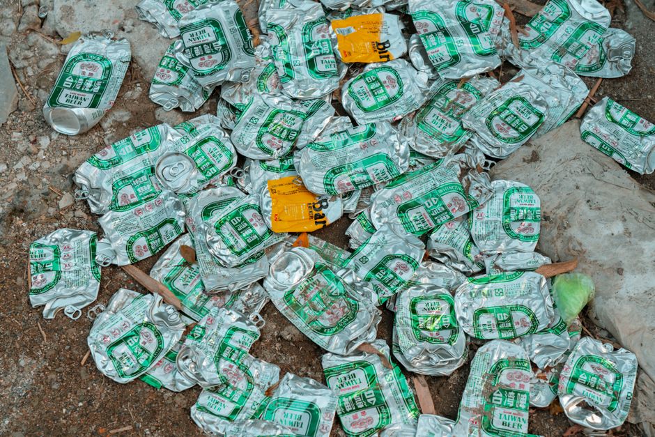 A collection of crushed aluminium cans scattered on a rough outdoor surface, primarily silver with green and red printed labels indicating they are Taiwanese beer cans. The cans vary in orientation, some lying flat while others are partially tilted or overlapping, showing signs of being emptied and flattened. Among the cans, there are small pieces of brown soil, dry leaves, and a few bits of plastic or paper packaging. In the background, there appears to be a white plastic bag partially buried beneath the debris, along with a small orange label or sticker attached to one of the cans. The scene is outdoors, suggesting an informal area for rubbish disposal, characteristic of waste accumulation that might require professional rubbish clearance from services like Rubbish Clearance Surrey, who handle such general waste and recyclables. The lighting is natural, highlighting the metal textures and worn exteriors of the cans, emphasizing the environmental context of littering and waste management.